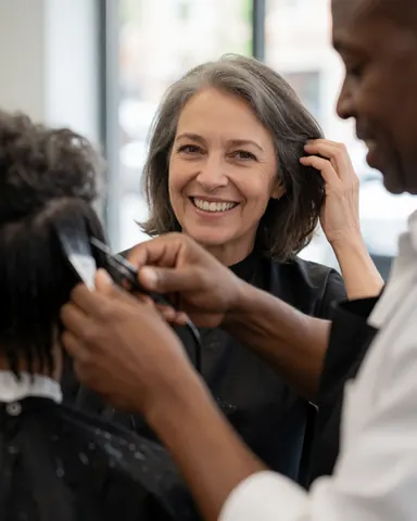 A hairdresser uses scissors and a comb to cut the hair of a smiling female client in a modern salon with natural light.