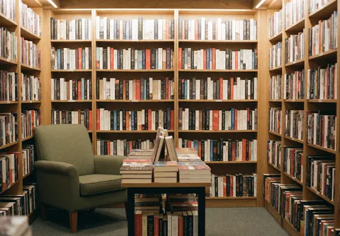 Warm bookstore interior featuring wooden bookshelves filled with books, a green chair, and a table with stacked books.