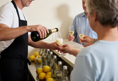 A bartender pours champagne into a glass for clients at a bar counter with lemons and bottles, capturing service in action at an event or wedding.