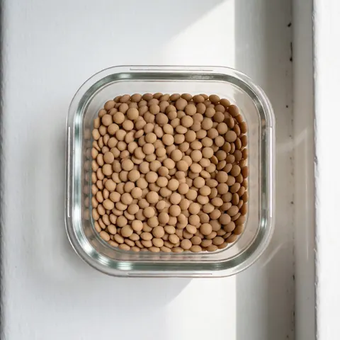 A clear glass jar filled with dry brown lentils sits on a white surface with natural lighting, suitable for nutrition and health recipes.