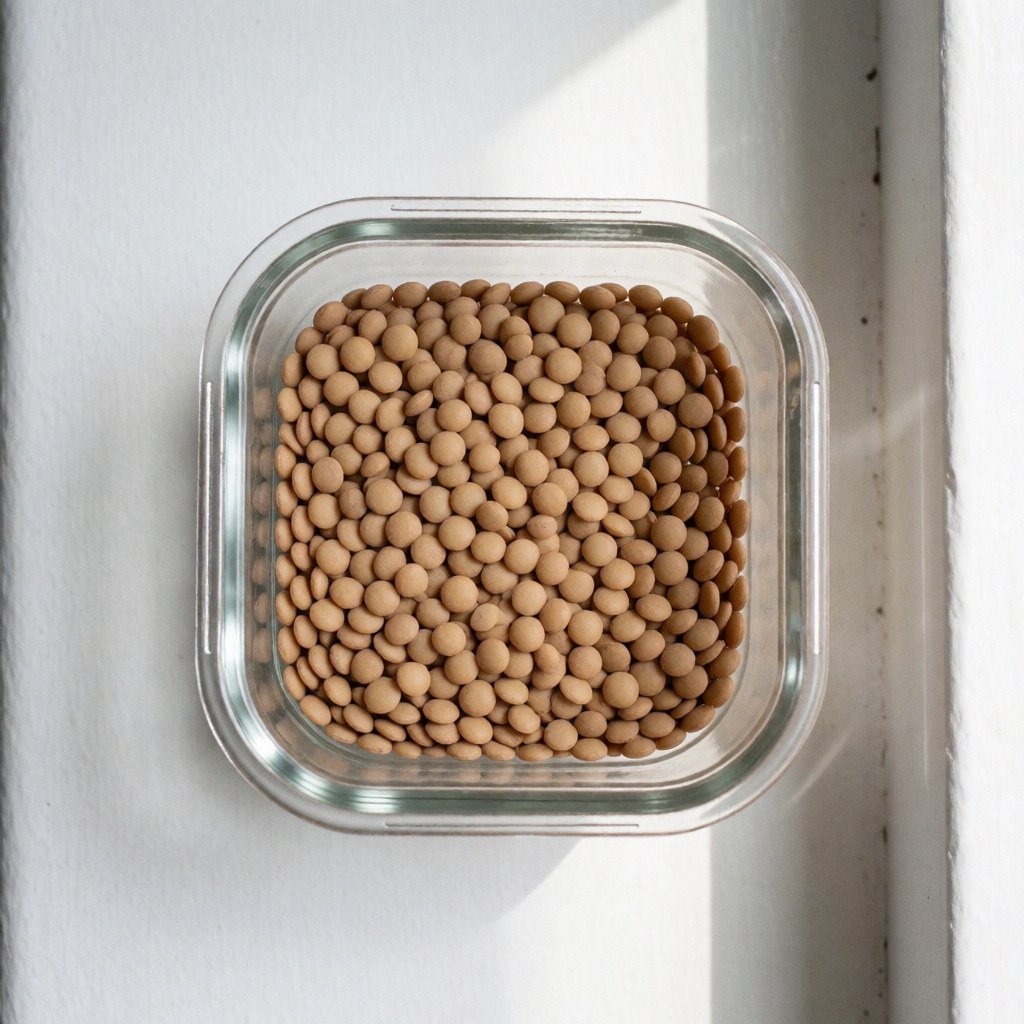 A clear glass jar filled with dry brown lentils sits on a white surface with natural lighting, suitable for nutrition and health recipes.