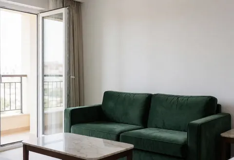 Elegant empty hotel suite featuring a green sofa, marble table, and large windows overlooking a balcony with natural light