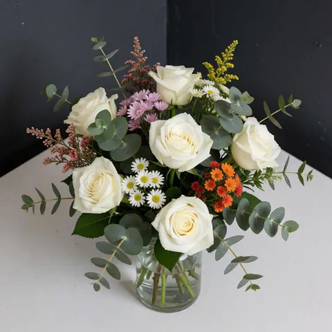 Elegant white rose and eucalyptus floral arrangement in a clear glass vase placed on a white table against a gray wall, perfect for weddings and events