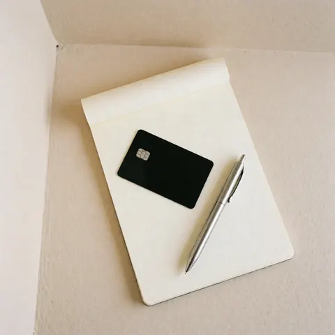 Black debit card and silver pen resting on a cream-colored notebook, set against a beige wall for a clean financial business product display