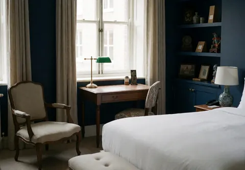 Empty hotel suite featuring a bed, chair, and desk with blue walls, natural light, and cultural decor details.