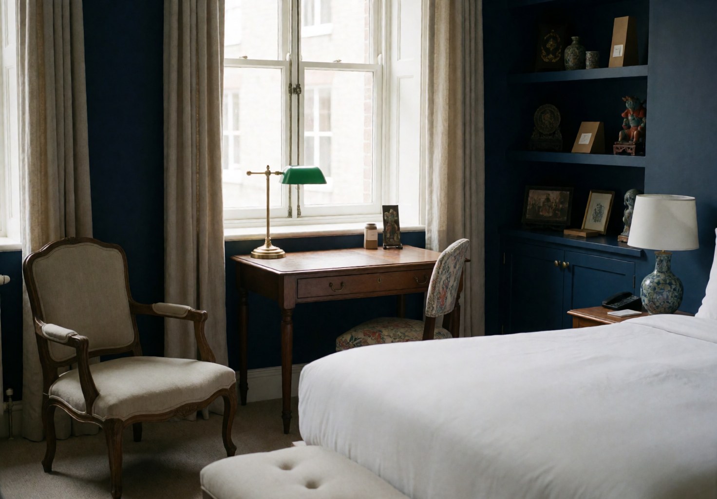Empty hotel suite featuring a bed, chair, and desk with blue walls, natural light, and cultural decor details.