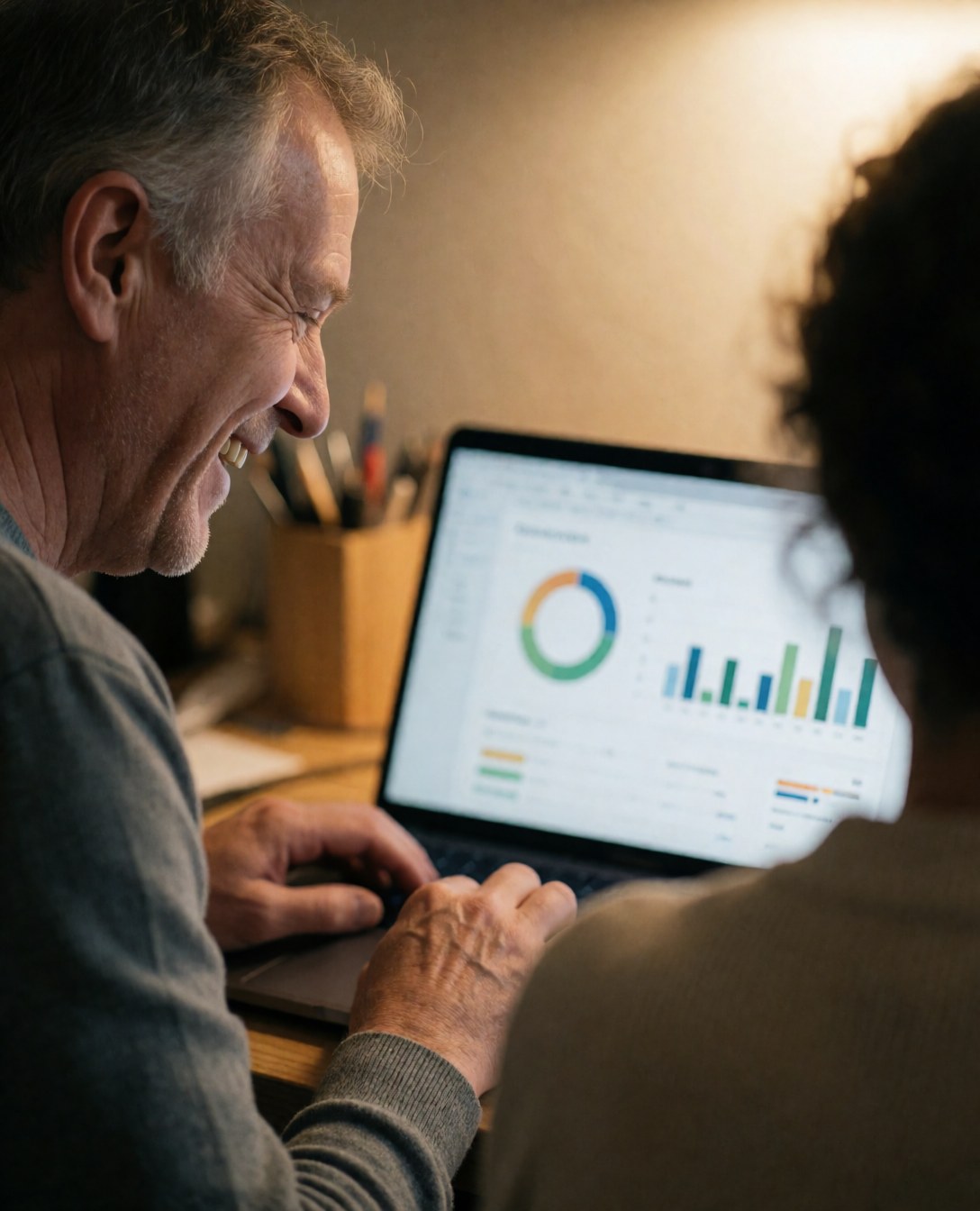 A smiling man reviews marketing data on a laptop in a warmly lit office with a colleague nearby.