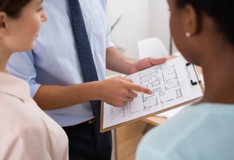 An advisor displays architectural blueprints to two attentive clients in a modern office, pointing out details during a professional team meeting.