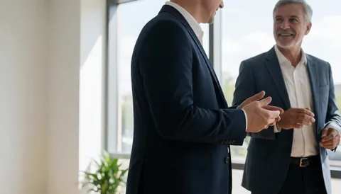 Two business men in suits and shirts conversing in a bright office corridor near windows and plants