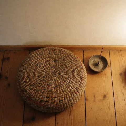 A woven meditation cushion and incense burner rest on a wooden floor against a white wall, creating a warm, relaxing atmosphere for mindfulness and wellness.