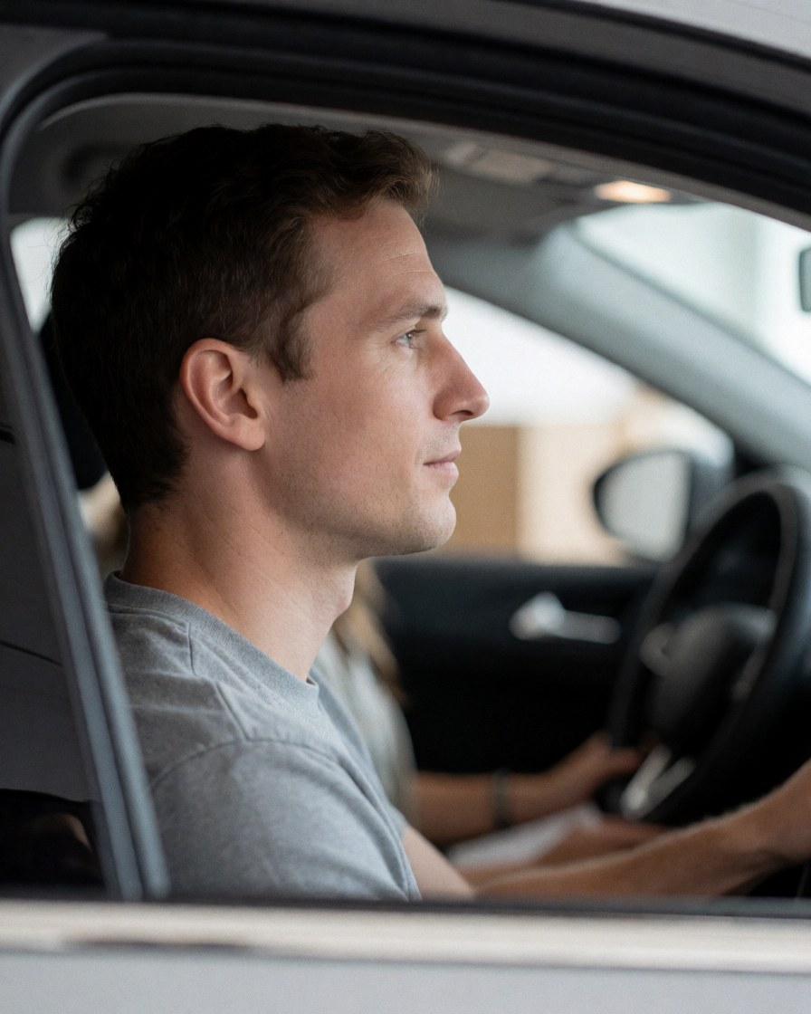 A driving school student sits in the driver's seat, looking ahead with concentration, hands on the steering wheel, with the car interior visible and a blurred background.
