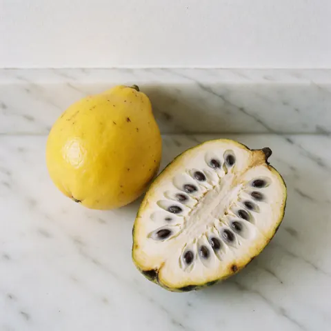 A whole lulo and a sliced soursop revealing white pulp and black seeds on a bright white marble counter with natural light.