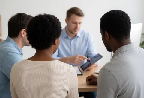 A professional insurance agent explains policy details to a seated couple in a modern office setting using a tablet.