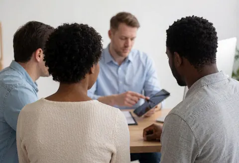 A professional insurance agent explains policy details to a seated couple in a modern office setting using a tablet.