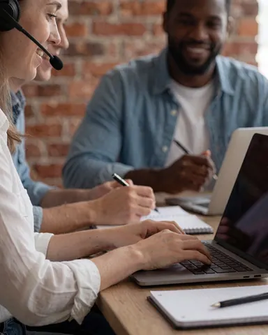 Man and woman collaborating in a virtual language class using a laptop, notebook, and pen in an office with a brick wall and natural light.
