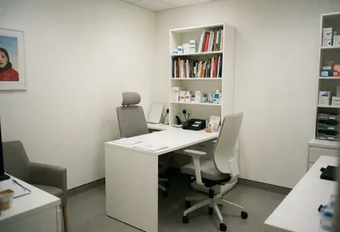 Modern medical office with white desk, ergonomic chairs, bookshelves with books and medications, neutral lighting, and a clean professional atmosphere.