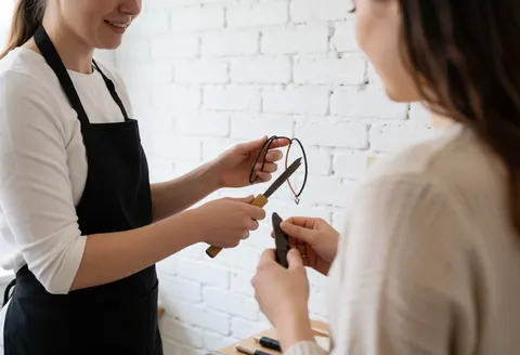 A saleswoman displays jewelry and accessories to a client in a clothing store with a white brick wall background and natural lighting