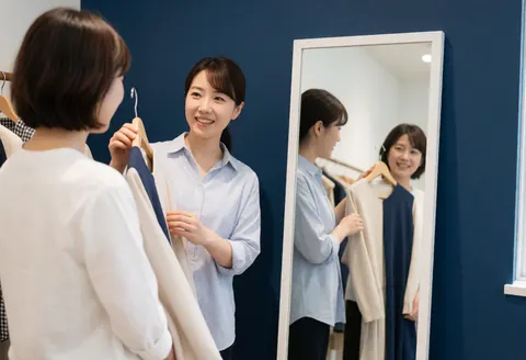 A stylist presents a garment to a smiling female client in front of a mirror inside a clothing store with blue walls and natural light.