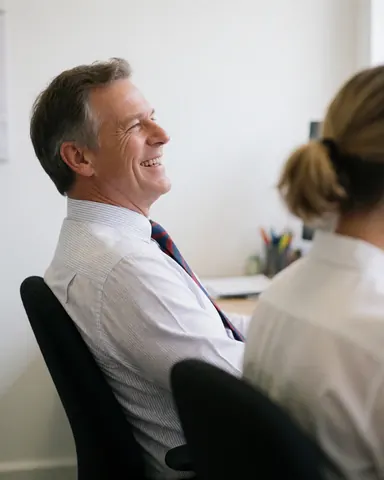 A smiling businessman in an office setting, seated on a chair, with a colleague standing behind him in a bright professional environment.