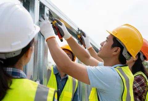 Workers wearing hard hats and safety vests assemble a metal framework outdoors using tools under a clear sky.