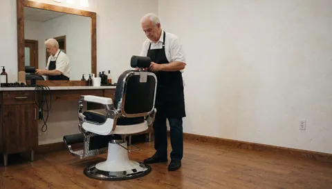 Senior barber adjusting a classic barber chair in an empty shop with white walls and wood floors, leaving space on the right for text or logo.
