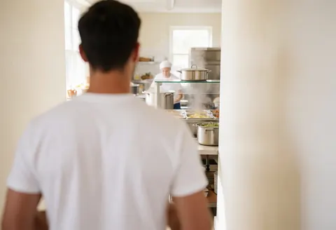 A volunteer walks through a bright community kitchen with a chef and steaming pots visible in the background.
