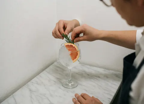 A hand garnishing a gin and tonic cocktail with a slice of grapefruit and a sprig of rosemary on a white marble surface with soft natural lighting.