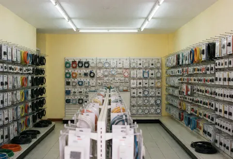 Well-organized electrician shop interior featuring shelves stocked with cables, switches, and electrical products against light yellow walls under fluorescent lighting.