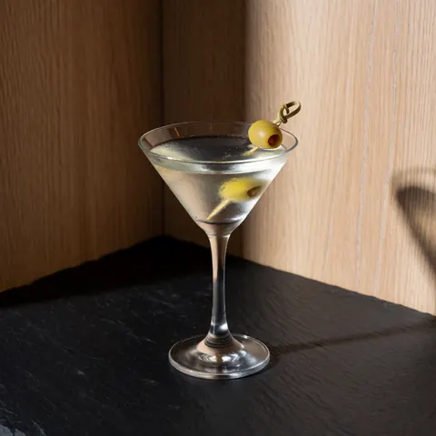 Elegant martini in a glass with an olive on a toothpick, placed on a dark slate surface against light wood walls with dramatic lighting.