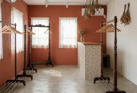 Clothing store interior featuring terracotta walls, polished concrete floors, vintage wooden hangers, and a mosaic counter with natural light from lace-curtained windows.