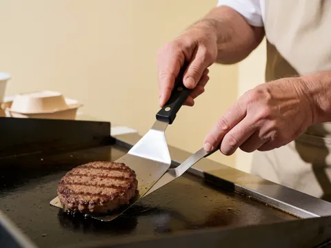 A close-up view of a juicy hamburger patty sizzling on a hot grill, with visible grill marks and steam rising from the cooking meat.