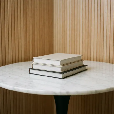A neat stack of hardcover books placed on a white marble table with wooden slats and a white wall background, ideal for bookstore product displays.