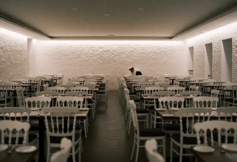 Empty event room featuring white tables and chairs against white brick walls with soft lighting and a person working in the background