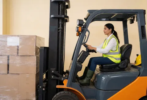 A woman in a reflective vest and yellow hard hat operates a forklift in an industrial warehouse, moving cargo boxes.