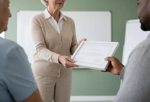 A female instructor hands a framed certificate to a student in a modern classroom setting, symbolizing language school achievement.