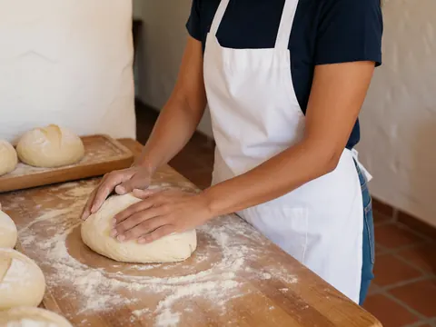 A woman shaping bread dough on a wooden table dusted with flour in a bakery with white walls and terracotta tile floor