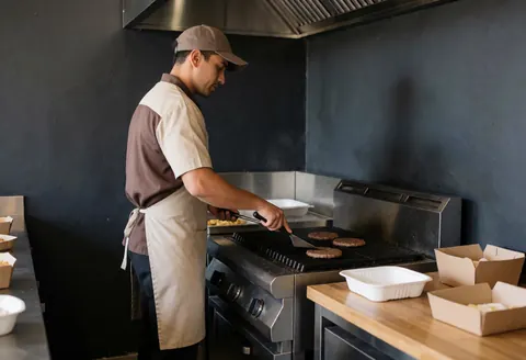 Professional chef flipping hamburgers on a stainless steel grill in an open kitchen with gray walls, capturing the fast food service in action.