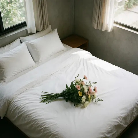 A neatly made bed with white linens and a vase of fresh flowers on a nightstand in a clean hotel room with concrete walls and natural light