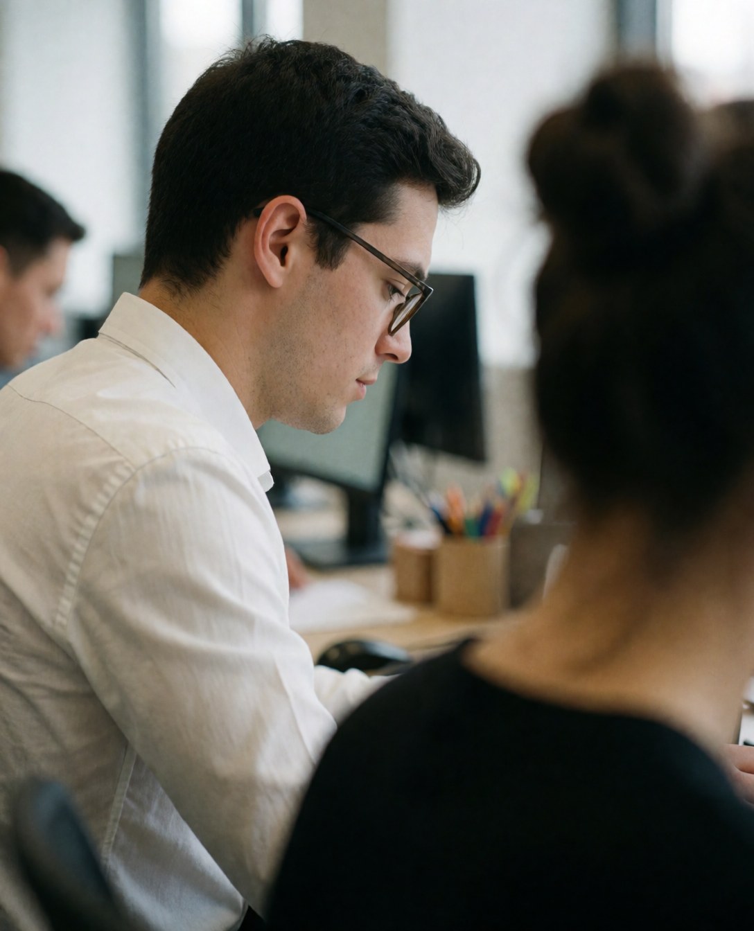 A young professional man wearing glasses and a white shirt sits at a modern office desk working on a laptop with a blurred background.