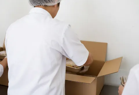 Worker packing food in a cardboard box, wearing gloves and a mask, in a clean and well-lit environment.