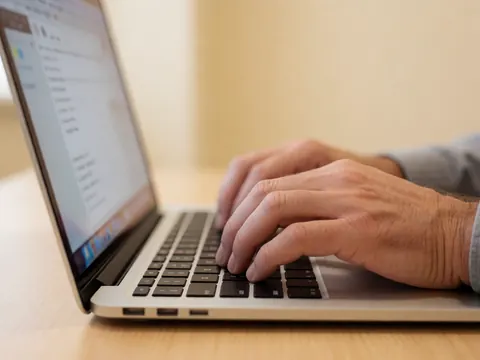 Close-up of an adult hand typing on a laptop keyboard placed on a wooden desk with a beige wall and warm lighting in the background.