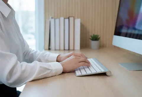 A professional person typing on a keyboard at a wooden desk with books and a plant in a modern office setting.