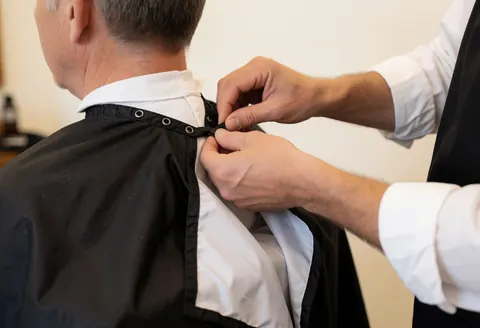 Close-up view of a barber's hands adjusting a protective cape around a client's neck in a barbershop setting