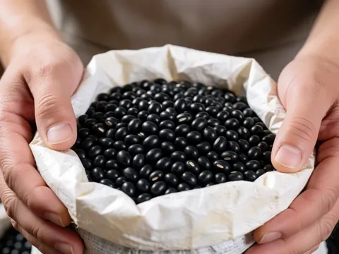 Close-up of hands holding a paper bag filled with black beans, highlighting the texture of the grains with natural lighting and a blurred background.