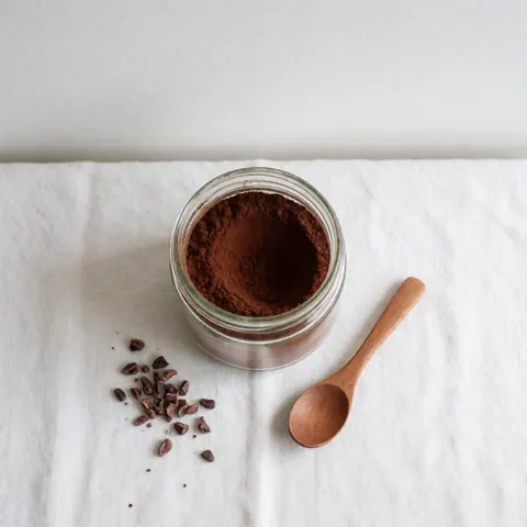 A glass jar filled with artisanal cocoa powder and a wooden spoon resting on a white background for product display