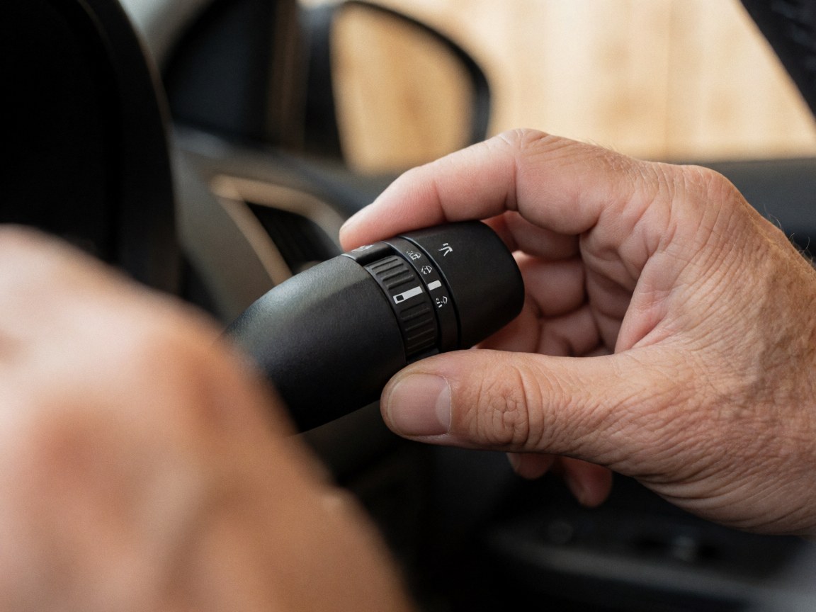 Close-up of an adult hand turning the headlight switch on the steering column with a blurred car interior background, suitable for driving school materials.