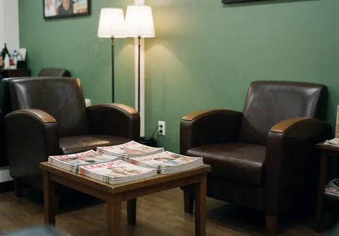 An empty waiting room in a barbershop featuring green chairs, a table, and magazines on the wall.