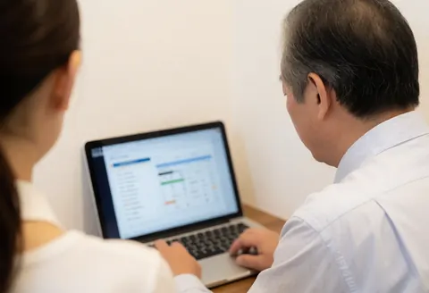 A business owner checks a cash flow account on a laptop while a woman stands beside him in a bright office with white walls and warm lighting.