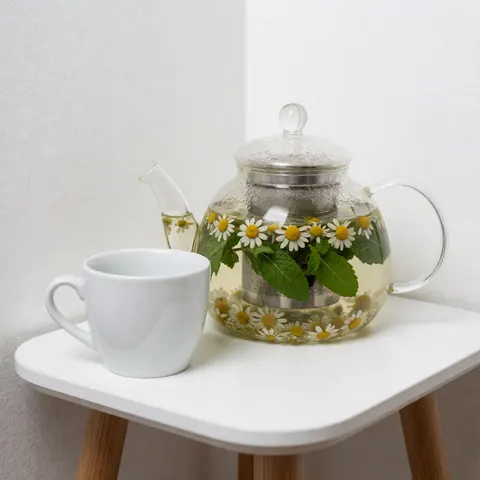 Glass teapot with chamomile and mint tea on white table and wall, creating a relaxing spa atmosphere
