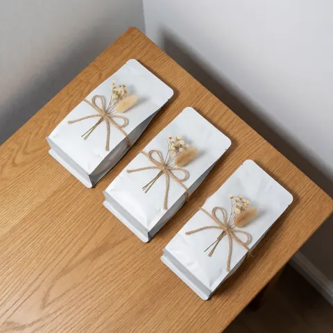 Three white coffee bags with natural twine and dried flowers arranged diagonally on a light wooden table with natural lighting and gray walls